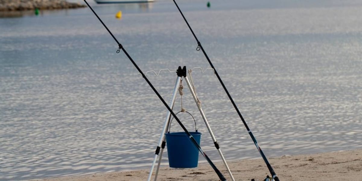 A flat-lay of sea fishing beach tripod accessories on the sand: spare head and cups, rod rests, sand spike and tray